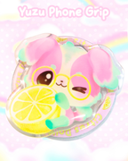 A pastel pink and mint green  phone grip featuring a cute cartoon dog character wearing glasses and holding a lemon slice, set inside a clear circular frame. The text 'Yuzu Phone Grip' is written at the top against a soft pink background with a faint rainbow.