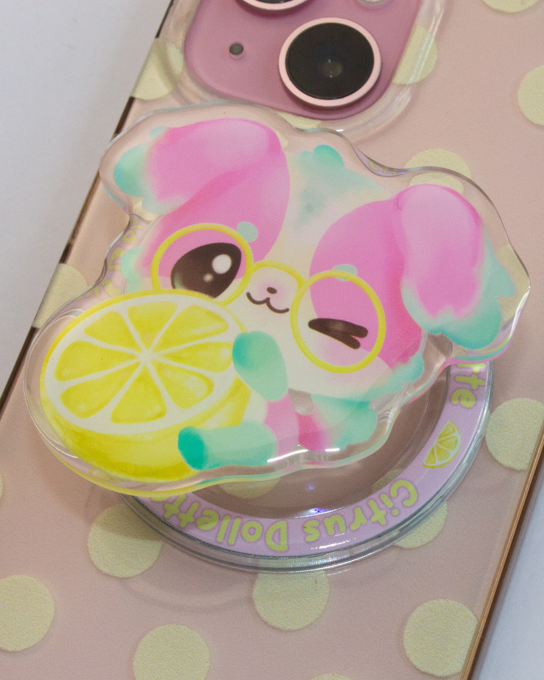 A close-up of a clear acrylic phone grip or stand featuring a pink cartoon dog character wearing yellow glasses and holding a yellow lemon slice, shown on a pale pink phone case with yellow polka dots.