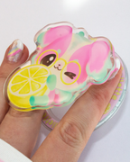 A hand holding a translucent acrylic charm shaped like a pink cartoon character wearing yellow glasses and holding a yellow lemon slice, with a matching round acrylic stand partially visible underneath.