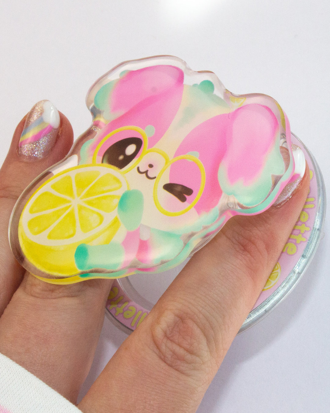 A hand holding a translucent acrylic charm shaped like a pink cartoon character wearing yellow glasses and holding a yellow lemon slice, with a matching round acrylic stand partially visible underneath.