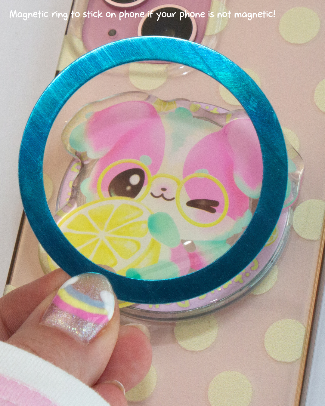 A hand with glittery rainbow nail polish holds a clear phone accessory with a bright blue magnetic ring. The accessory is shown over a phone case featuring a pink cartoon dog with yellow glasses and a lemon slice graphic.