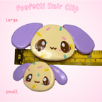 Two cartoonish hair clips with purple ears and yellow faces on a pink background, with a ruler for scale.
